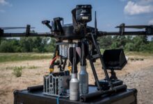 A Skylab Shoolika Mk6 drone is seen propped up on top of a black box in a gravelly, grassy area. The drone is painted black, with some of its wirings exposed. Its rotary blades are folded inwards. The background is a tree-covered area, with the ble sky filling in the far back.