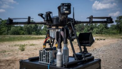 A Skylab Shoolika Mk6 drone is seen propped up on top of a black box in a gravelly, grassy area. The drone is painted black, with some of its wirings exposed. Its rotary blades are folded inwards. The background is a tree-covered area, with the ble sky filling in the far back.