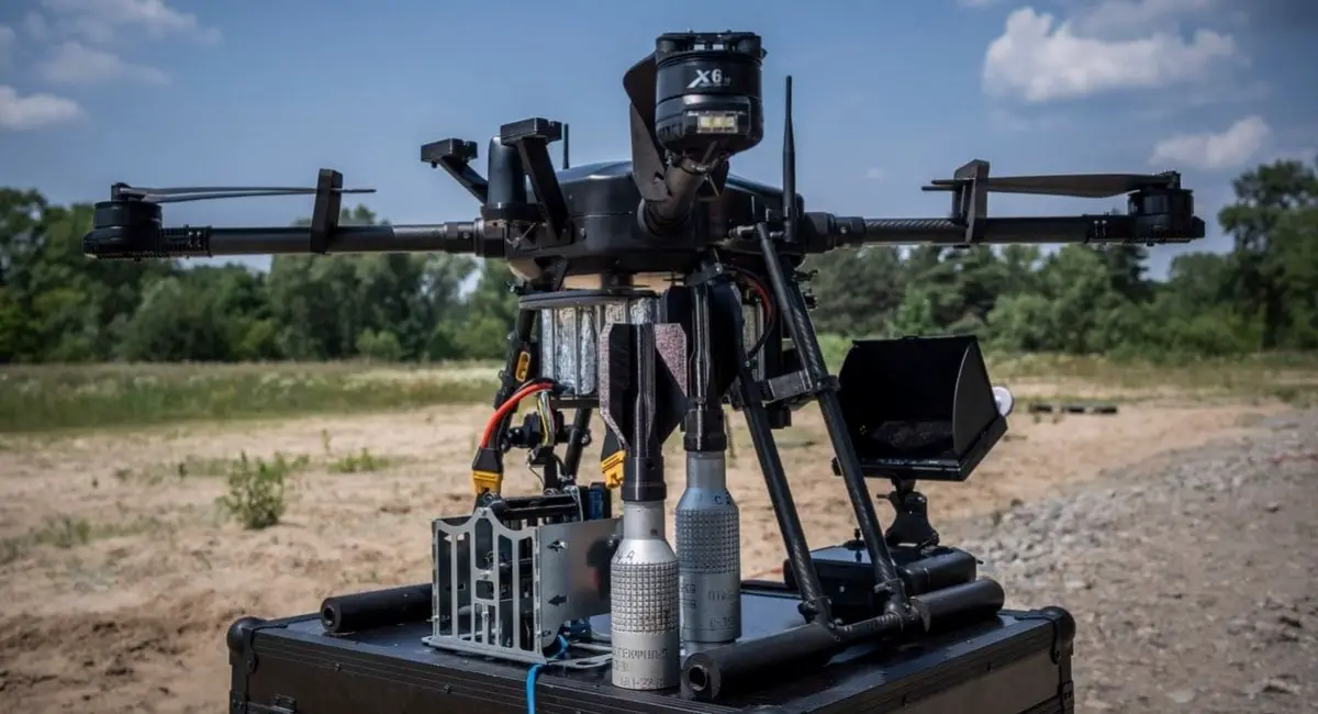 A Skylab Shoolika Mk6 drone is seen propped up on top of a black box in a gravelly, grassy area. The drone is painted black, with some of its wirings exposed. Its rotary blades are folded inwards. The background is a tree-covered area, with the ble sky filling in the far back.