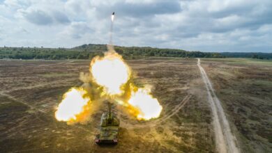 A Panzerhaubitze 2000 tank is seen firing a 155mm ammunition at the Ravlunda test field in Sweden. Three balls of blazing fire are seen jutting out of the tank's gun barrel, with the ammunition flying off upwards. The test field is a greenish-brown plains dotted with small plant growths. In the distant background, a hilly landscape covers the horizon. The blue sky is full of light-gray clouds.