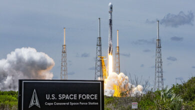 A rocket launches from at Cape Canaveral Space Force Station, Florida. On the left of the foreground, a sign that says "U.S. SPAC FORCE Cape Canaveral Space Force Station" is seen. The Space Force Logo is on the left of the sign.