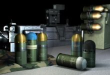 A computer-generated image displays Rheinmetall's line of 40mm x 53 ammunition. Ten ammos are seen on the image: three are standing upright on the left opposite two on the right, one is laid down in front of the latter, and four are attached to a metallic-looking box on the right side of the background. A long, gray grenade launcher is seen displayed farther in the background.