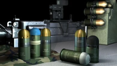 A computer-generated image displays Rheinmetall's line of 40mm x 53 ammunition. Ten ammos are seen on the image: three are standing upright on the left opposite two on the right, one is laid down in front of the latter, and four are attached to a metallic-looking box on the right side of the background. A long, gray grenade launcher is seen displayed farther in the background.