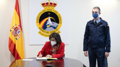 Spanish Defense Minister Margarita Robles is seen signing a document inside what appears to be a conference room. An office personnel is seen standing by her left. The Spanish Air and Space Force emblem is displayed on the background, and the Spanish flag is positioned to her right.