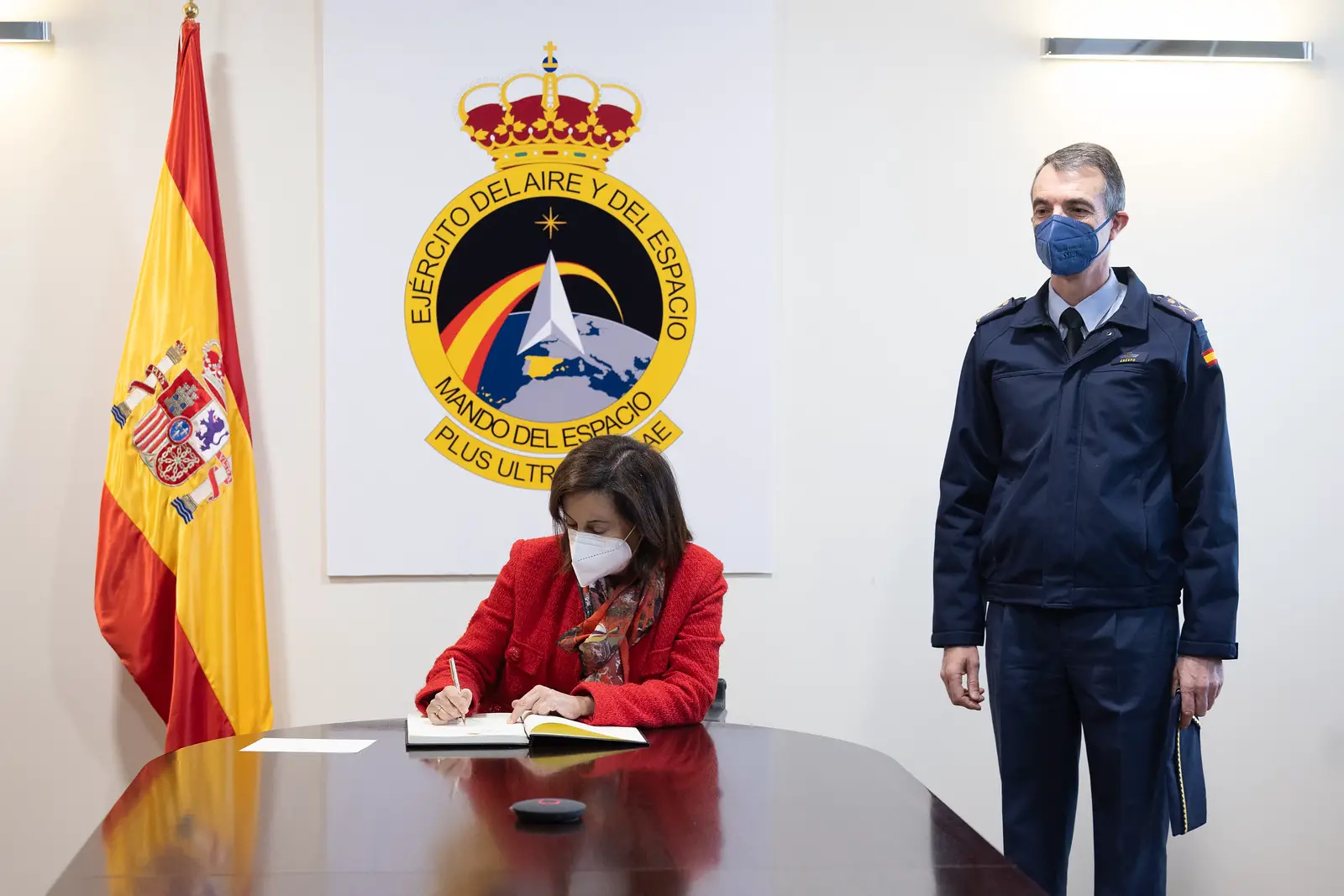 Spanish Defense Minister Margarita Robles is seen signing a document inside what appears to be a conference room. An office personnel is seen standing by her left. The Spanish Air and Space Force emblem is displayed on the background, and the Spanish flag is positioned to her right.