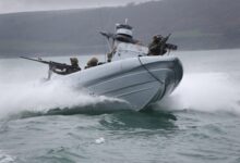 The Commando Raiding Craft is seen in action in Plymouth Sound. It's seen moving in a coastal body of water, with at least six personnel on the craft. Two soldiers are manning what appears to be machine guns, one in front of the boat and the other in the back. In the background, a coastal land mass is seen amid foggy weather.
