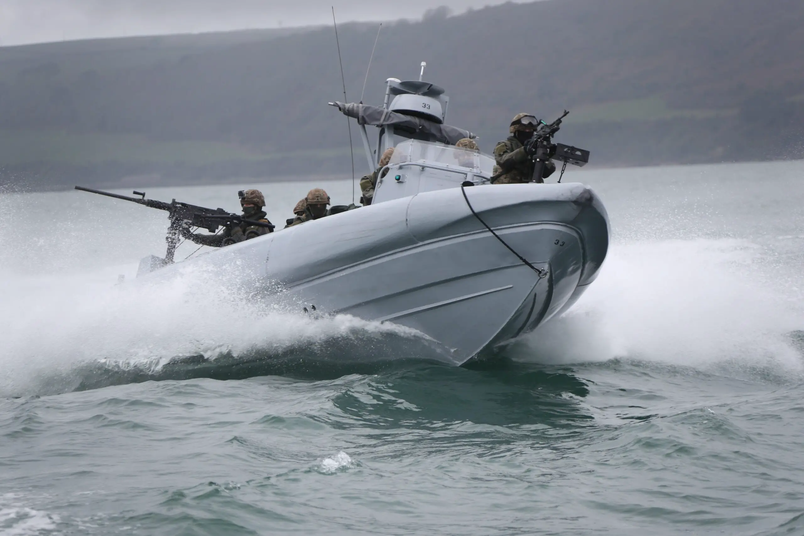 The Commando Raiding Craft is seen in action in Plymouth Sound. It's seen moving in a coastal body of water, with at least six personnel on the craft. Two soldiers are manning what appears to be machine guns, one in front of the boat and the other in the back. In the background, a coastal land mass is seen amid foggy weather.