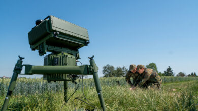A Thales surveillance system is seen propped up in a grassy plains. It's positioned on the left side of the foreground, with two military personnel in camouflage on the far right in the background. The sky is a clear, cloudless blue.
