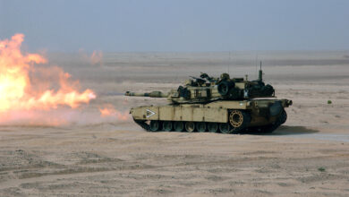 An Abrams M1A1 tank is seen initiating live fire in a desert field. Red-orange flames appear to be coming out of its turret. The tank's hull blends well with the area's dry, sandy environment. The background is a dim blue sky.