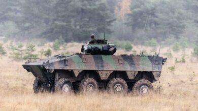 The French Army's VBCI Infantry Fighting Vehicle is seen moving across a brown, grassy area. The vehicle is painted in green, black, and brown camouflage. The background is a forested area.