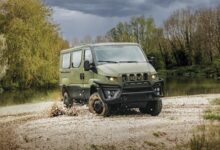 An Iveco Military Utility Vehicle is seen traversing a flooded and muddy terrain. The camouflage-green van's wheels are covered in mud, and the brown water on the ground is seen rippling and splashing because of the vehicle. The area appears to be a swamp or a forest, with overgrown bushes and trees in the background. The sky is dark and cloudy.