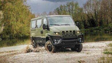 An Iveco Military Utility Vehicle is seen traversing a flooded and muddy terrain. The camouflage-green van's wheels are covered in mud, and the brown water on the ground is seen rippling and splashing because of the vehicle. The area appears to be a swamp or a forest, with overgrown bushes and trees in the background. The sky is dark and cloudy.