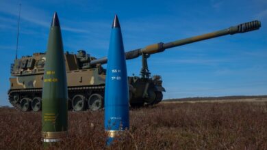 Two 155mm artillery ammunition are placed upright in a grassy field. The one on the left has its shell painted green, and the other is paited blue. A camouflage-painted battle tank is stationed behind them. The background is a clear blue sky.