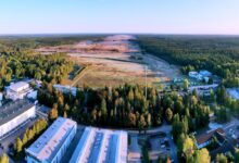 A view of the landscape where Werk Niedersachsen will be constructed. The foreground of the image is a complex made up of several buildings. Behind them, a forested area is shown, its middle stripped off of trees and its brown land flattened. The area stretches over the horizon.