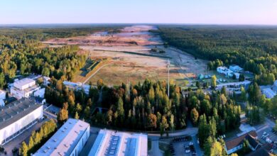 A view of the landscape where Werk Niedersachsen will be constructed. The foreground of the image is a complex made up of several buildings. Behind them, a forested area is shown, its middle stripped off of trees and its brown land flattened. The area stretches over the horizon.