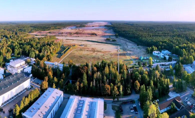 A view of the landscape where Werk Niedersachsen will be constructed. The foreground of the image is a complex made up of several buildings. Behind them, a forested area is shown, its middle stripped off of trees and its brown land flattened. The area stretches over the horizon.