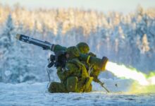 Two camouflaged soldiers fire off a Javelin anti-tank missile pointing northwest. The launcher is seen emitting yellow-green flames from its back. The scene is taking place in a snowy area, with a forest of snow-covered trees blurred out in the back.