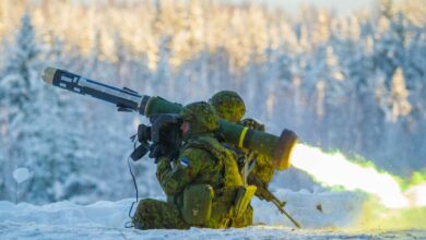Two camouflaged soldiers fire off a Javelin anti-tank missile pointing northwest. The launcher is seen emitting yellow-green flames from its back. The scene is taking place in a snowy area, with a forest of snow-covered trees blurred out in the back.