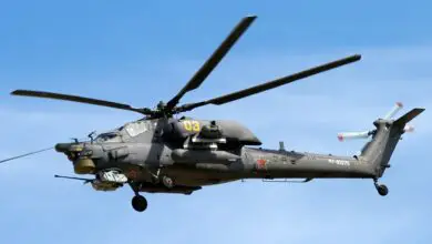 A Mil Mi-28 helicopter is seen hovering on the clear blue sky. On its gray-green hull, the number "03" is painted in yellow, as well as two red stars and the unit name "RF-93275" in white. Some characters on the Russian alphabet are also painted in black.