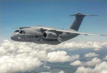 An Embraer C-390 Millennium aircraft is seen flying above the clouds. The gray aircraft has the words "HUNGARIAN AIR FORCE" painted on the side of the cockpit in black. The number 610 is also painted on the jet engine, the wing, and the side of the cockpit, just below one of the side windows. The background is a bright blue sky.