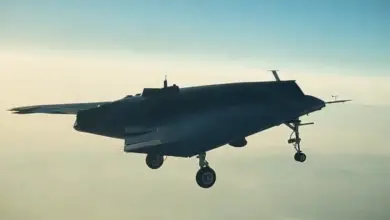 A gray Turkish Aerospace Industries Anka-3 unmanned combat aerial vehicle is seen flying above the clouds. The warplane is largely flat-shaped, with its four landing wheels jutting out underneath. The background is a light blue sky, with white clouds reflecting the sun's orange rays below.