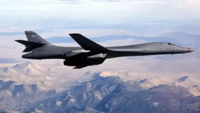 A black B-1B Lancer bomber is seen flying above a mountainous region. The mountains' land have colors ranging from forest's greens to desert yellows and whites. The sky above is cloudy and white.