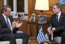 Greek Prime Minister Kyriakos Mitsotakis is seen speaking with US Secretary of State Anthony Blinken. They' re sat across each other, with a table in the middle decorated with the Greek and US flags.