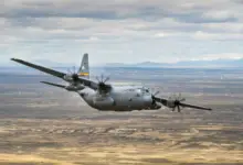A C-130 Hercules flies over a sandy plains. The plane is colored gray, with a yellow stripe on its tail. It has "US AIR FORCE" painted on its side in black, as well as "WYOMING AIR GUARD", below its wing. The background is the landscape; a wide stretch of land occupies most of the space, with green grass and yellow-white sands scattered evenly. Over the horizon, a mountain range is seen. Some of its peaks are snowy and white. The sky above is full of thin, long clouds.