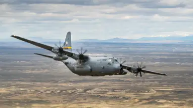 A C-130 Hercules flies over a sandy plains. The plane is colored gray, with a yellow stripe on its tail. It has "US AIR FORCE" painted on its side in black, as well as "WYOMING AIR GUARD", below its wing. The background is the landscape; a wide stretch of land occupies most of the space, with green grass and yellow-white sands scattered evenly. Over the horizon, a mountain range is seen. Some of its peaks are snowy and white. The sky above is full of thin, long clouds.