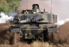 The British Army's Challenger 3 Main Battle Tank is seen moving across a dusty grassland. The tank is painted in black and green camouflage, and its tracked wheels are shrouded in brown dust from the ground. In the back, white smoke is seen, presumably from the tank's exhaust.