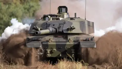 The British Army's Challenger 3 Main Battle Tank is seen moving across a dusty grassland. The tank is painted in black and green camouflage, and its tracked wheels are shrouded in brown dust from the ground. In the back, white smoke is seen, presumably from the tank's exhaust.
