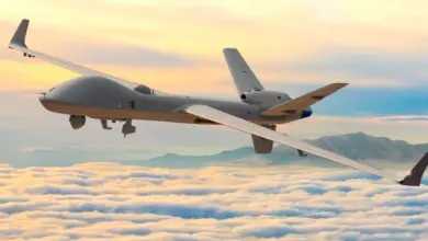 A gray General Atomics MQ-9B SkyGuardian is seen flying above the clouds. On the far right side of the background, a mountain range is seen peeking out. The scenery is tinged in the setting sun's orange hues.