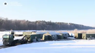 Hospitainer's Role II Hospital, a mobile, inflatable building, is seen set up in a snowy field outside a forested area in the background. Two container trucks are parked outside of it, and two cargo truck containers are also positioned on the far right of the field.