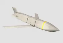A Lockheed Martin Joint Air-to-Surface Standoff Missile is seen front and center of the image. The background is plain white. The missile has fixed wings and a tail, painted dirty white with two black stripes and one yellow stripe painted on its body.