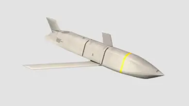 A Lockheed Martin Joint Air-to-Surface Standoff Missile is seen front and center of the image. The background is plain white. The missile has fixed wings and a tail, painted dirty white with two black stripes and one yellow stripe painted on its body.