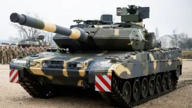 A camouflage-painted Leopard 2A7HU tank is seen stationed in a sandy area. Another tank is parked on the far left side of the background, with camouflage-wearing soldiers standing in attention in front of it. Some trees are seen poking out the far back of the image. The sky is a dull, foggy white.