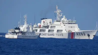 A white Philippine Coast Guard ship is stationed in the waters of the West Philippine Sea, facing a larger Chinese Coast Guard ship. The sea is a deep blue color, and the sky is a dull blue.