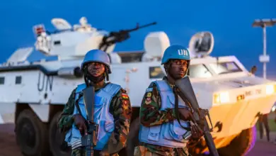 Two uniformed soldiers stand front and center of the image, staring straight. They both hold automatic rifles, pointed down. They are wearing a blue helmets and vests. The soldier on the right has 'UN' written in white in front of his helmet. Underneath their vests, they have a camouflage uniform on. A white tank with 'UN' written on its body in black is seen stationed in the background. Its orange headlights are on. The background is a dark blue sky.