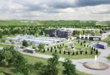 An artist's rendering of Rheinmetall's Várpalota plant. The complex shows a roofed parking lot three buildings, and what appears to be a small park. Three trucks and a bus are seen parked by the perimeter of the site. A roundabout is also shown on the lower right corner of the image. Most of the surrounding area is dotted by trees and grass.