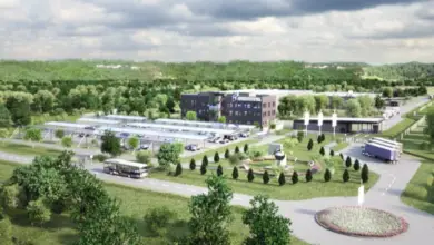 An artist's rendering of Rheinmetall's Várpalota plant. The complex shows a roofed parking lot three buildings, and what appears to be a small park. Three trucks and a bus are seen parked by the perimeter of the site. A roundabout is also shown on the lower right corner of the image. Most of the surrounding area is dotted by trees and grass.