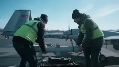 Two individuals are carrying a Desert Sting 16 bomb, preparing to install it under the wing of a Bayraktar TB2 drone. The individuals are wearing neon-green high-visibility vests. Both the bomb and the drone are painted gray. They appear to be set outside a hangar. The background is a clear blue sky with some thin white clouds.