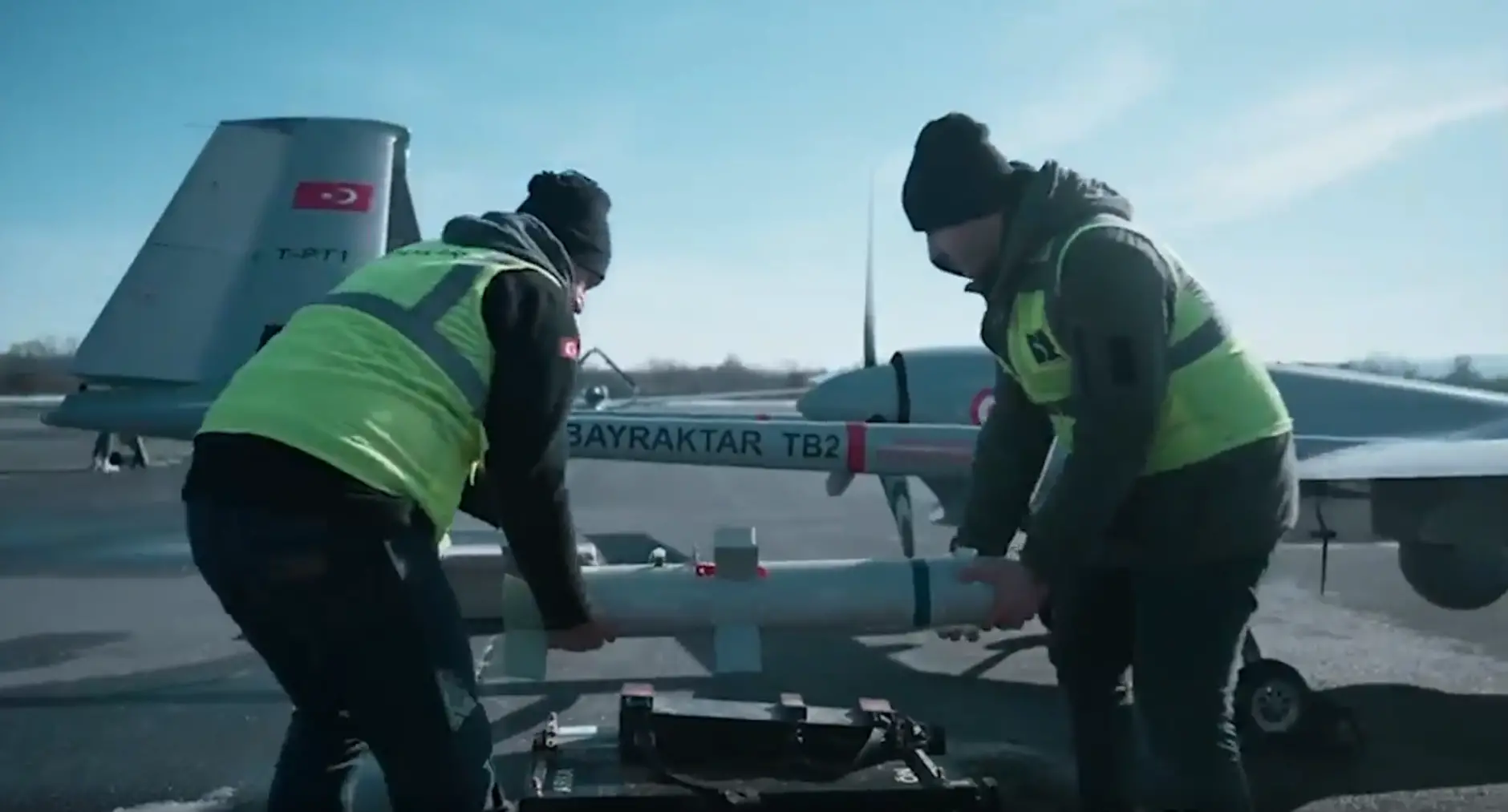 Two individuals are carrying a Desert Sting 16 bomb, preparing to install it under the wing of a Bayraktar TB2 drone. The individuals are wearing neon-green high-visibility vests. Both the bomb and the drone are painted gray. They appear to be set outside a hangar. The background is a clear blue sky with some thin white clouds.