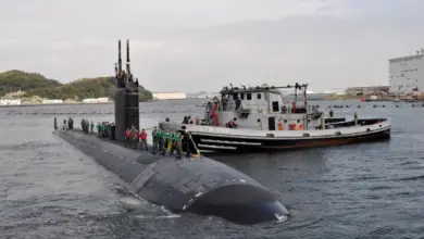 The USS Columbus submarine is seen partially submerged in the waters of what appears to be a pier. A boat with "YTB 798" painted on its side is seen right beside the black submarine. On top of its hull, around 25 people are seen standing, most of them wearing lifevests. In the background, a mountainous area is seen over the left of the horizon. On the right, two buildings that appear to be warehouses are also visible.