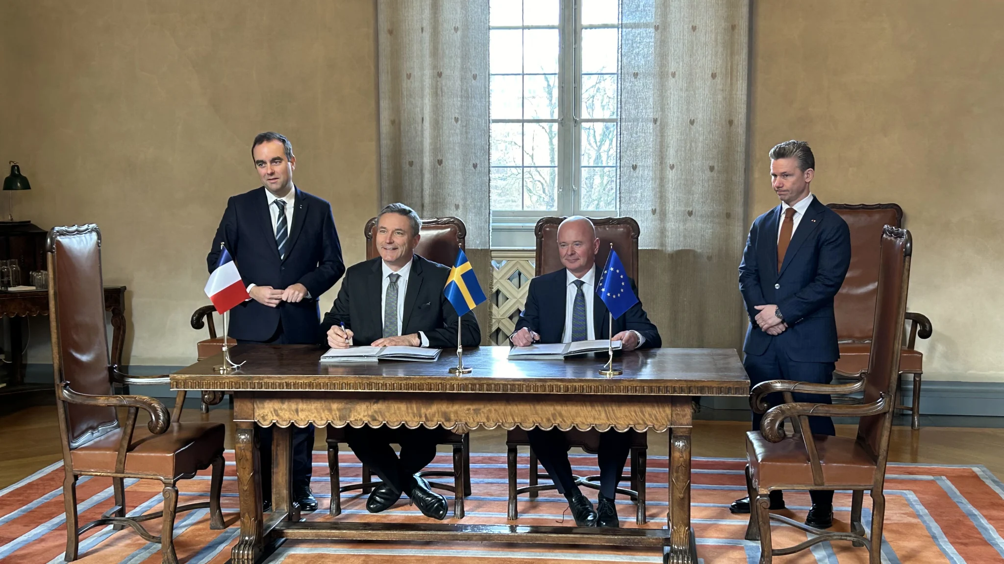 MBDA CEO Éric Béranger and Saab CEO Micael Johansson are seen signing a letter of intent on a table decorated with France, Sweden, and the EU's flags. On the left, they are witnessed by French Minister for the Armed Forces Sébastien Lecornu, and on the right, Swedish Minister for Defence Pål Jonson.