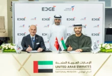 Turkish Aerospace President and CEO Professor Temel Kotil and EDGE Group Platforms & Systems President Khaled Al Zaabi are seen signing documents during a memorandum of agreement ceremony. In the back, a facilitator oversees the procedure. A white panel with EDGE and Turkish Aerospace's logos printed on it serves as their background. The table where the signing is taking place on has the UAE, Turkey, and the company's flags propped up on it.