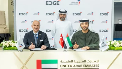 Turkish Aerospace President and CEO Professor Temel Kotil and EDGE Group Platforms & Systems President Khaled Al Zaabi are seen signing documents during a memorandum of agreement ceremony. In the back, a facilitator oversees the procedure. A white panel with EDGE and Turkish Aerospace's logos printed on it serves as their background. The table where the signing is taking place on has the UAE, Turkey, and the company's flags propped up on it.