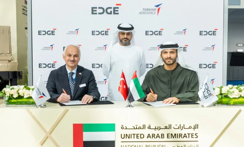 Turkish Aerospace President and CEO Professor Temel Kotil and EDGE Group Platforms & Systems President Khaled Al Zaabi are seen signing documents during a memorandum of agreement ceremony. In the back, a facilitator oversees the procedure. A white panel with EDGE and Turkish Aerospace's logos printed on it serves as their background. The table where the signing is taking place on has the UAE, Turkey, and the company's flags propped up on it.