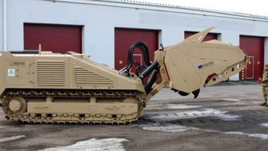A DOK-ING MV-10 mine-clearing vehicle can be seen outside of a warehouse. The brown-painted armored vehicle has tracked wheels and a tiller mechanism on its front. On the right, a camouflaged soldier is seen standing and looking at it.