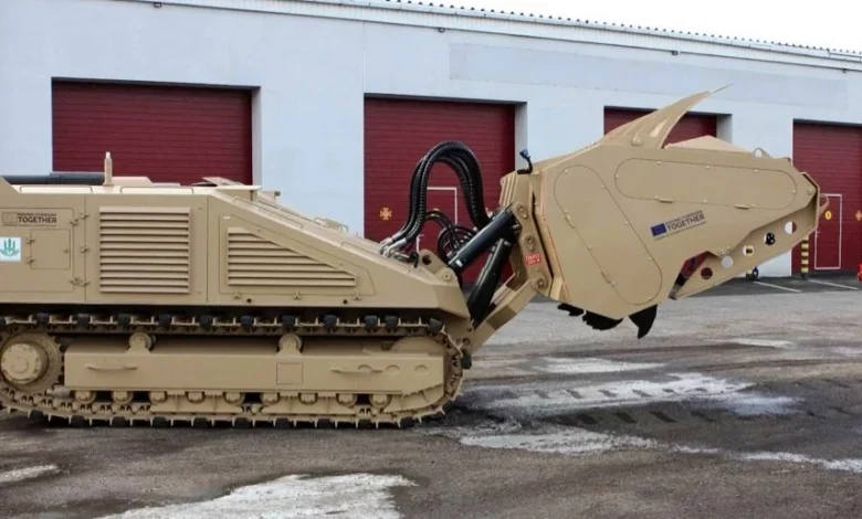 A DOK-ING MV-10 mine-clearing vehicle can be seen outside of a warehouse. The brown-painted armored vehicle has tracked wheels and a tiller mechanism on its front. On the right, a camouflaged soldier is seen standing and looking at it.