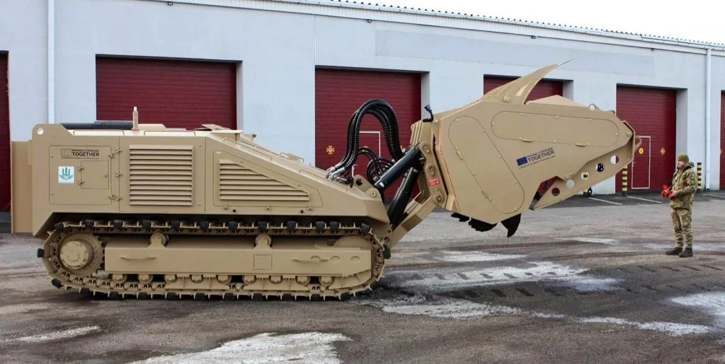 A DOK-ING MV-10 mine-clearing vehicle can be seen outside of a warehouse. The brown-painted armored vehicle has tracked wheels and a tiller mechanism on its front. On the right, a camouflaged soldier is seen standing and looking at it.